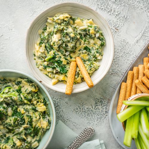 Slow cooker spinach artichoke dip with kale in three bowls with celery and breadsticks.