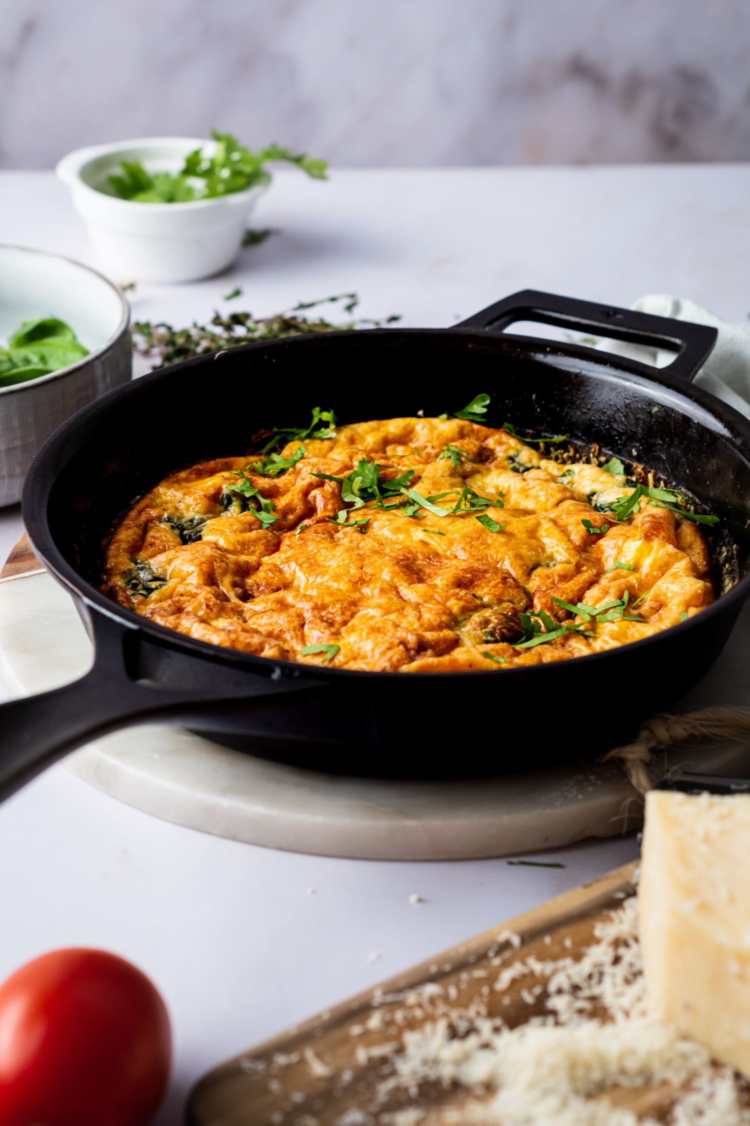 Spinach and cheese frittata in a skillet with tomatoes, herbs, and Parmesan cheese on the side.