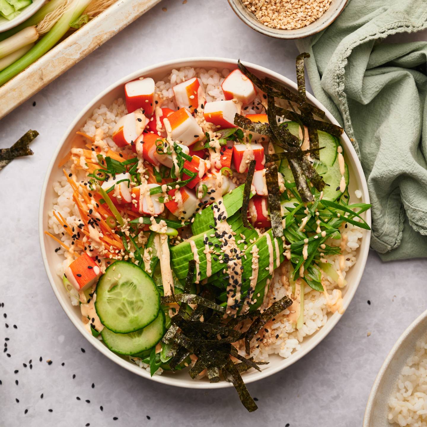 Sushi bowl with imitation crab, sushi rice, nori seaweed strips, cucumbers, carrots, and spicy mayo in a white bowl.