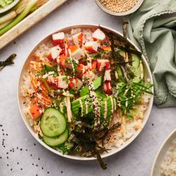 Sushi bowl with imitation crab, sushi rice, nori seaweed strips, cucumbers, carrots, and spicy mayo in a white bowl.