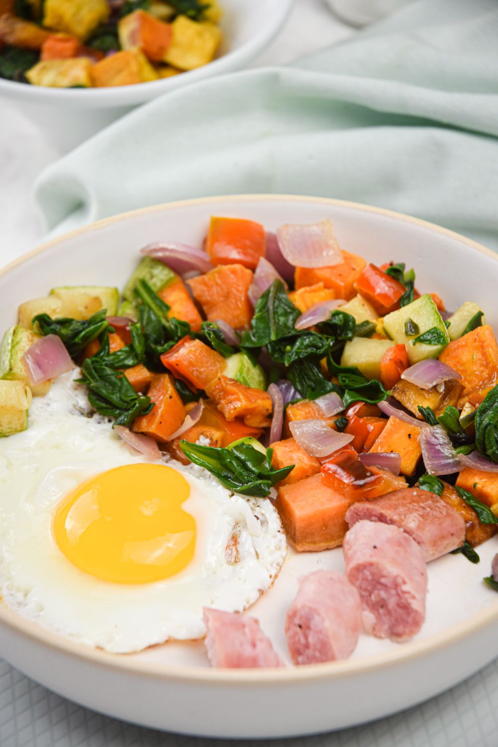 Sweet potato breakfast hash in a bowl with vegetables, a fried egg, and sliced sausage.