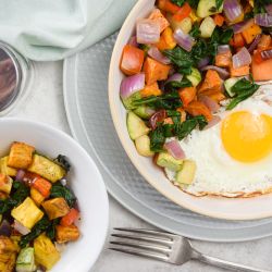 Sweet potato hash with sweet potatoes, zucchini, onions, peppers, and a fried egg on a plate with sliced sausage.