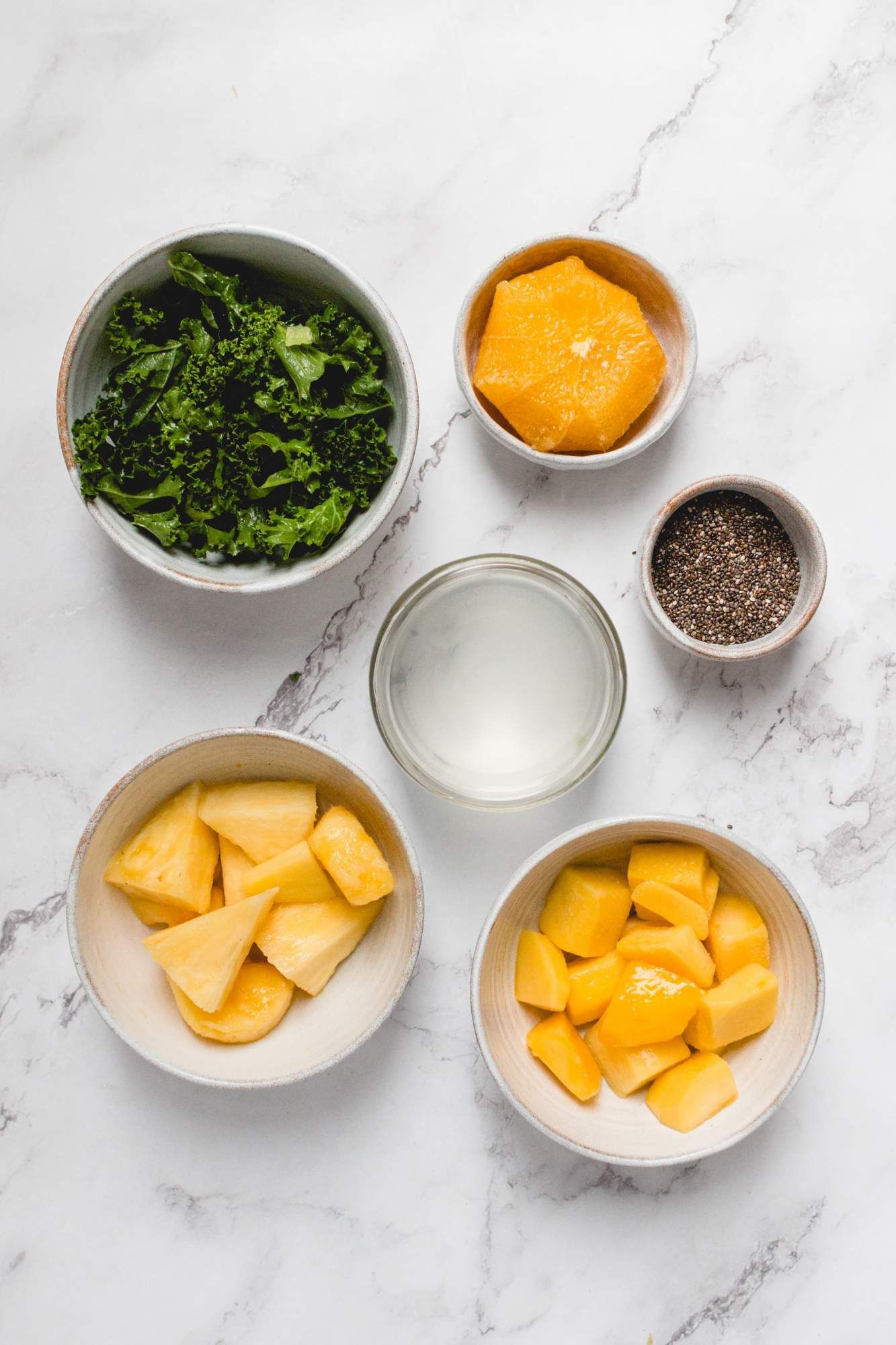 Ingredients for a kale smoothie including fresh kale, pineapple, mango, coconut water, oranges, and chia seeds.