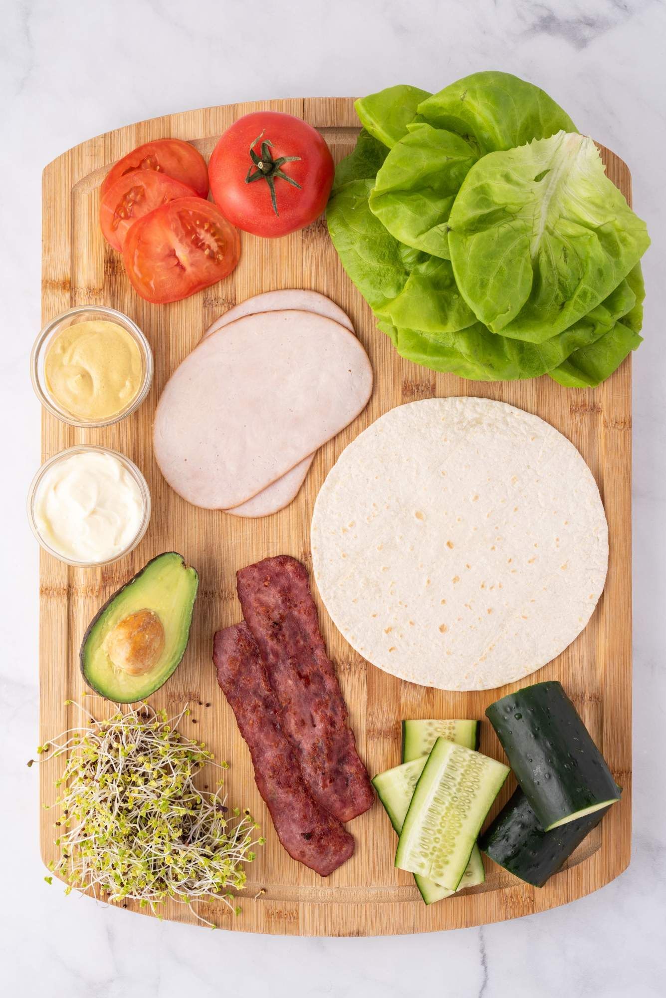 Ingredients for a turkey wrap with deli turkey, bacon, avocado, wraps, vegetables, and sauce.