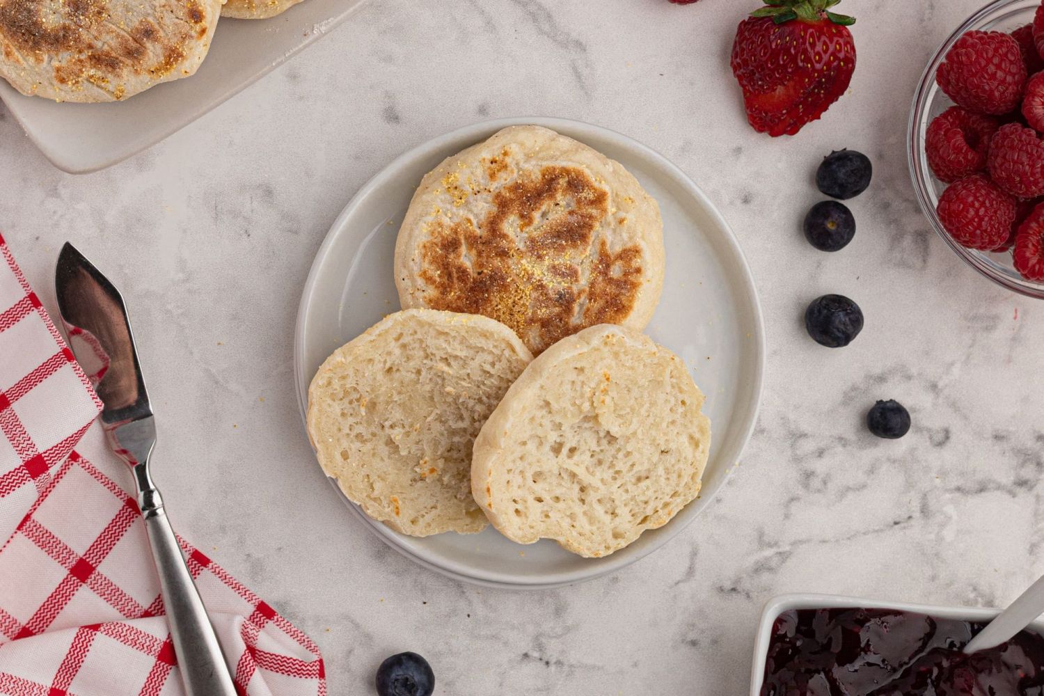 Toasted homemade English muffins with cornmeal cut in half with jam and berries on the side.