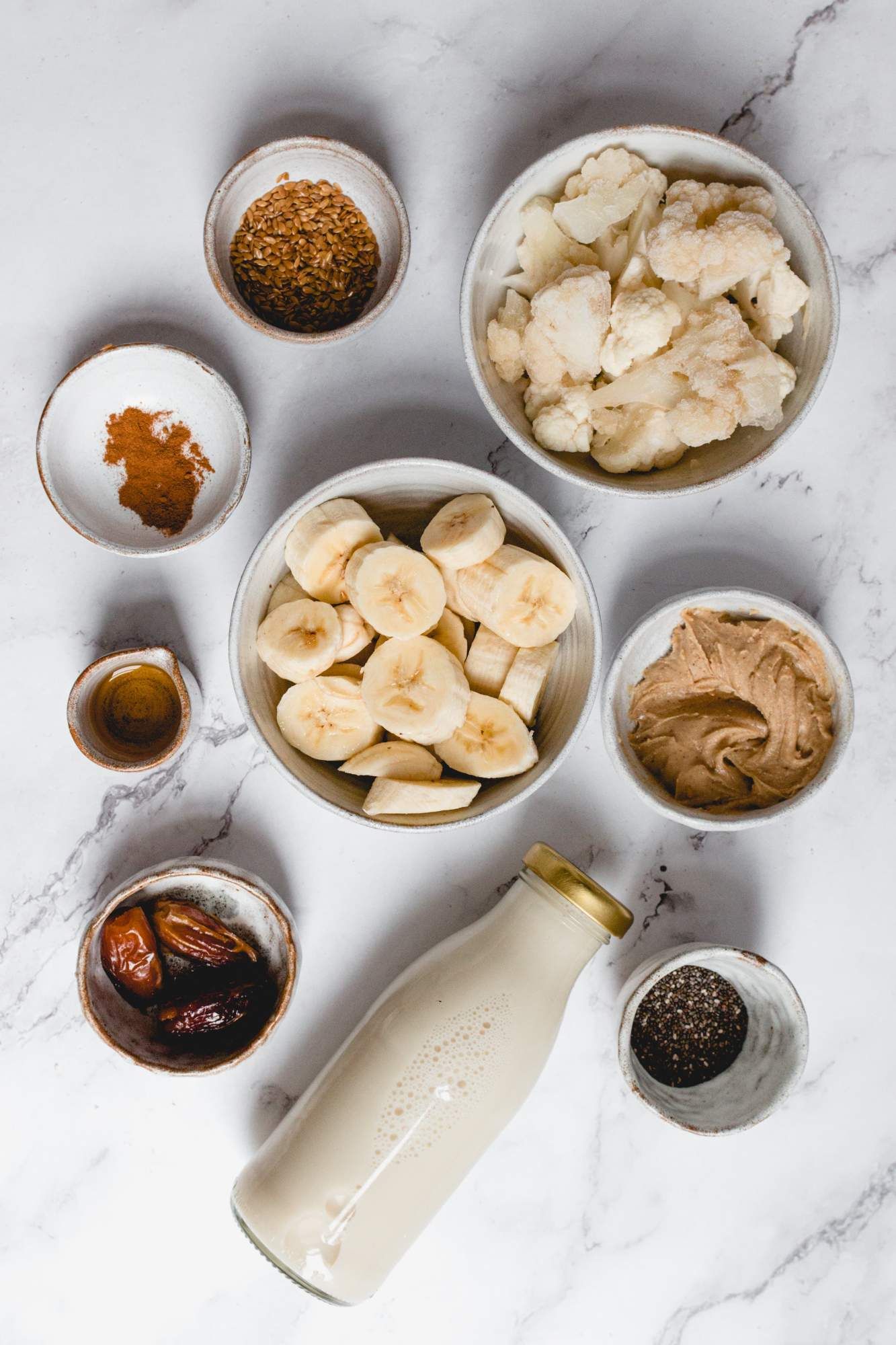 Ingredients for a vanilla cinnamon protein shake including bananas, cauliflower, almond butter, cinnamon, and seeds.